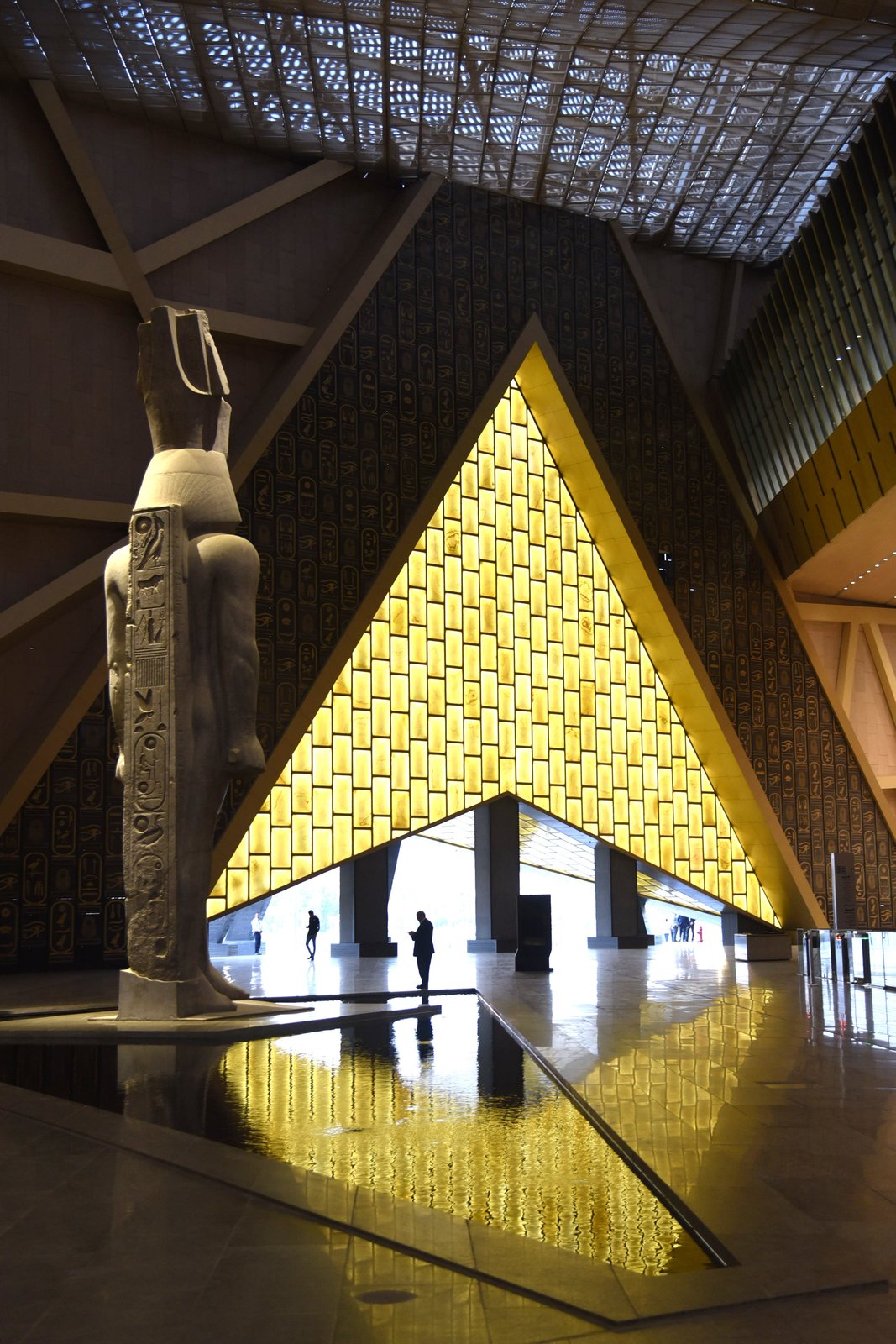 The museum’s monumental entrance atrium unfolds as a transitional space between the desert light and the built interior. At midday, sunlight filters through translucent surfaces and reflects across the floor, creating a dynamic play of golden tones that shifts throughout the day.  
This carefully designed effect establishes a symbolic dialogue between the Egyptian sun and the architectural materiality, turning the atrium into a space of welcome and contemplation.